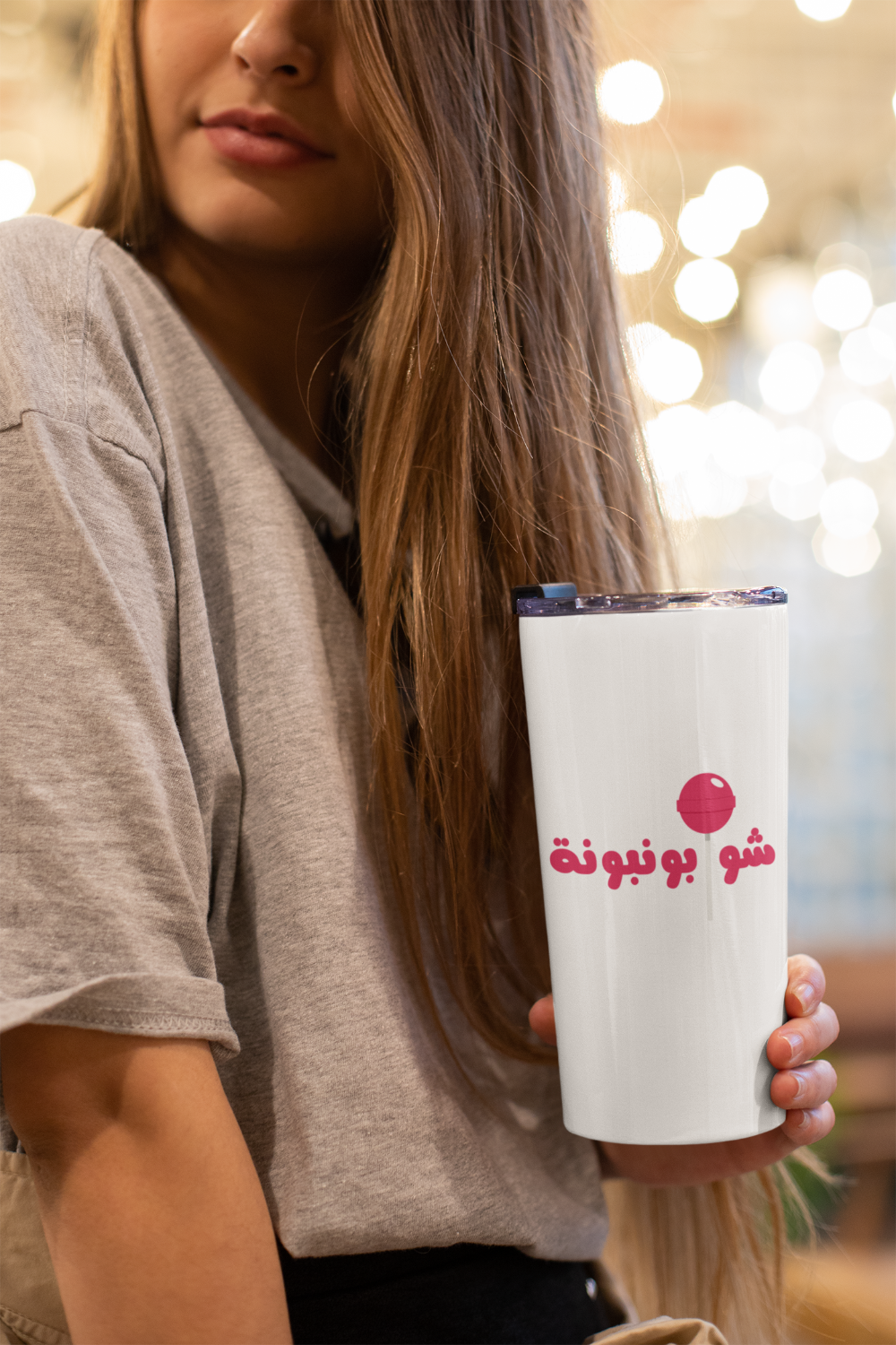 Person holding a white tumbler with Arabic text and a logo, blurred lights in the background