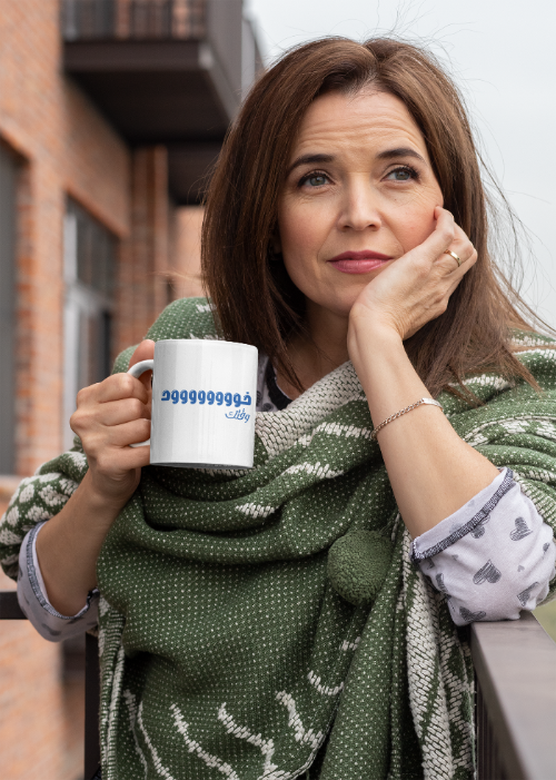 Woman holding a mug with text, wrapped in a green scarf, standing outdoors.