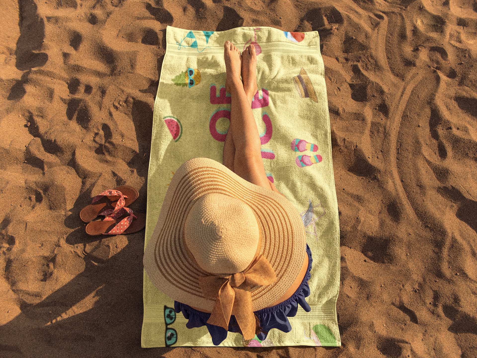 Person lying on a green beach towel with summer icons, wearing a wide straw hat, with sandals placed on the sand.