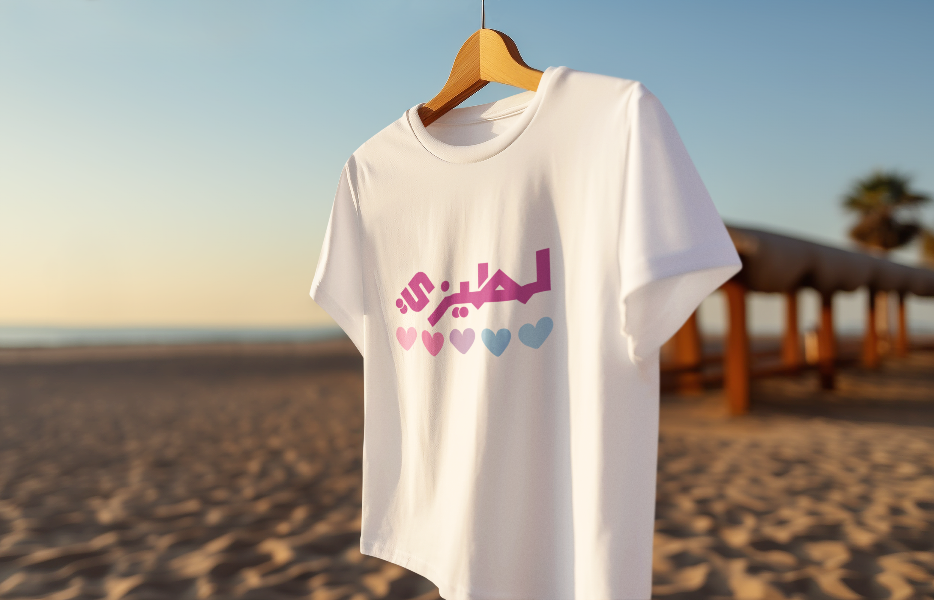 White t-shirt with colorful text and heart designs on a wooden hanger against a beach backdrop.