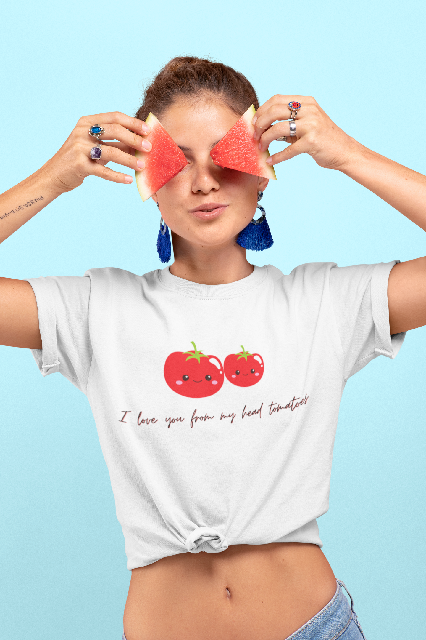 Woman wearing a white t-shirt with tomato design and text "I love you from my head tomatoes", holding watermelon slices over her eyes against a light blue background.